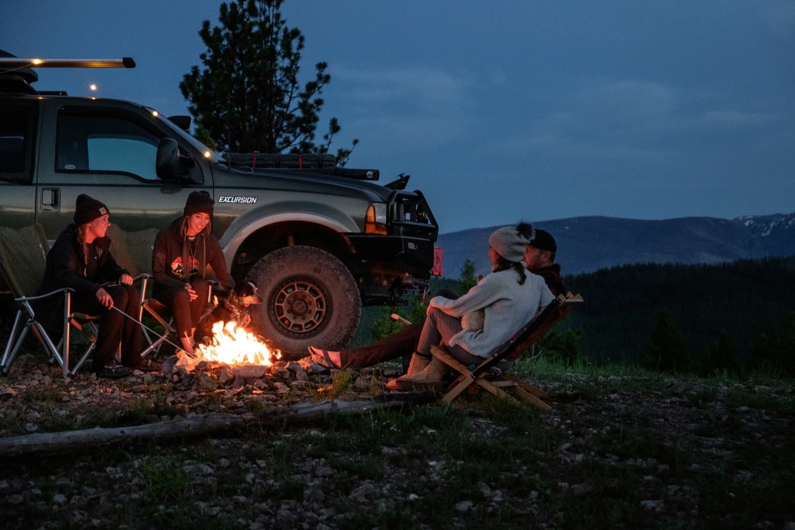 Four overlanders sitting around a campfire at dusk next to a green Ford Excursion with mountains in the background