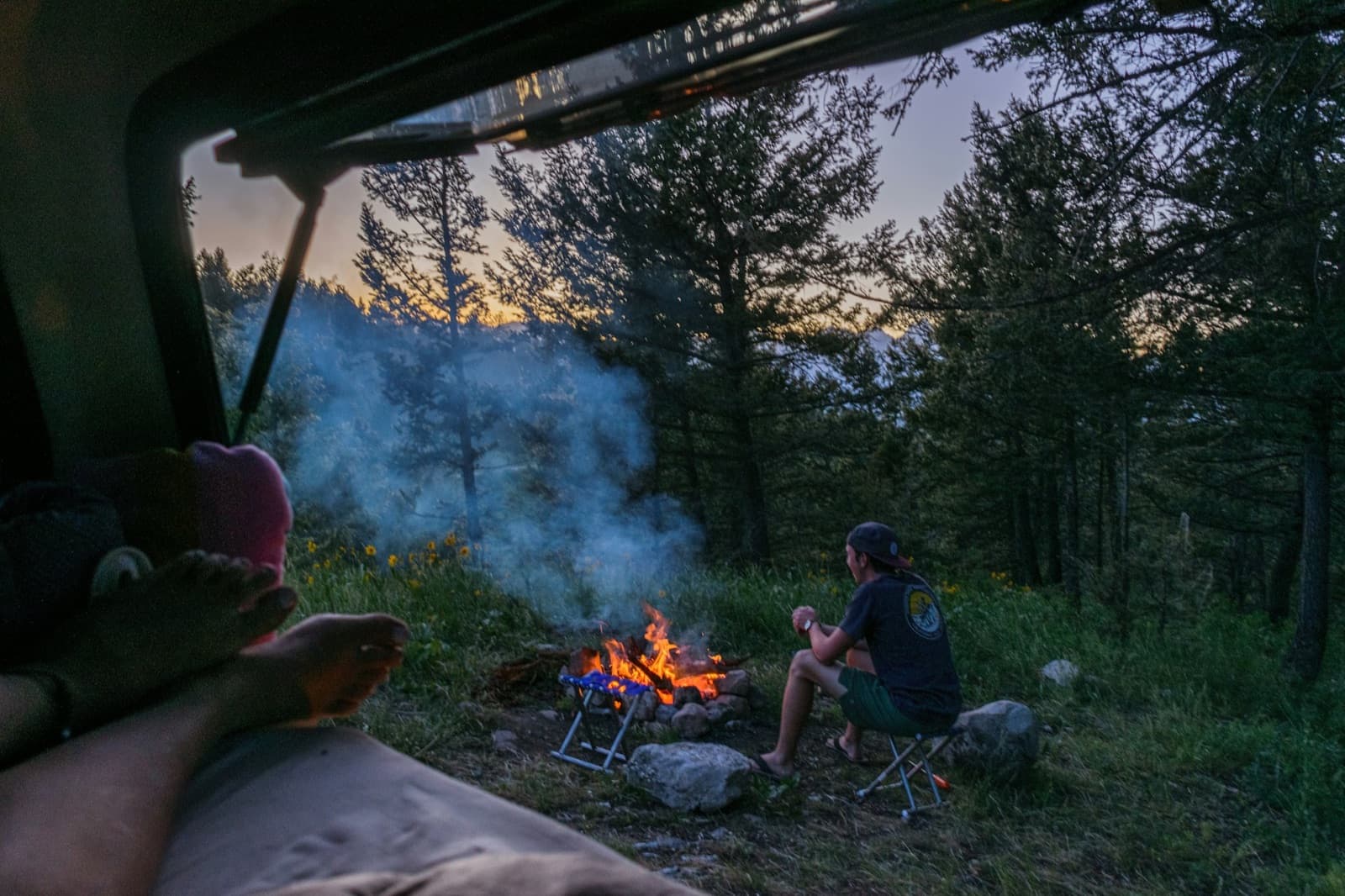 POV from inside an overland rig looking out at a campfire with a person sitting beside it at dusk in a pine forest